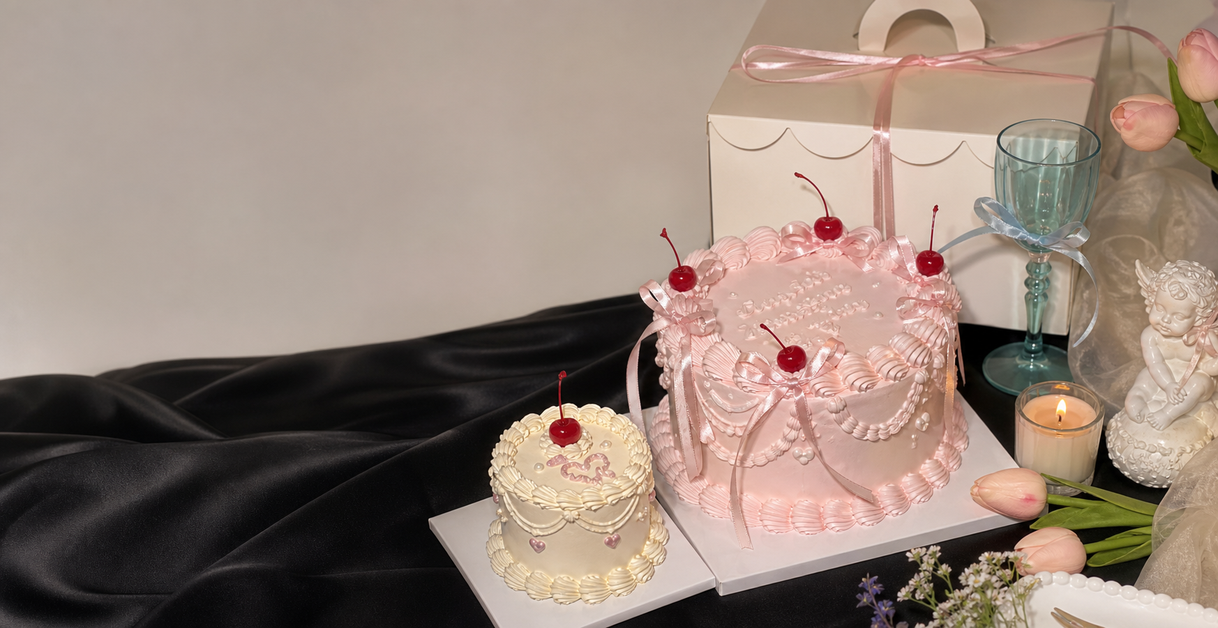 Pastel pink and cream custom cakes with ribbons, cherries, a cream cake box, tulips, glassware, and candlelight.